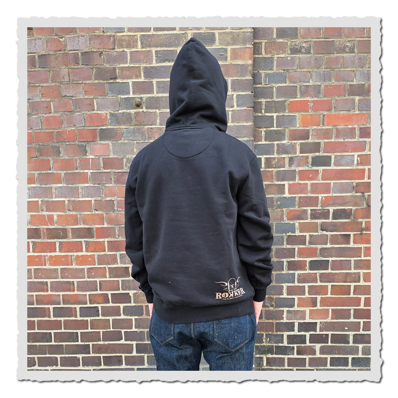 Rückansicht einer Person mit schwarzem Kapuzenpullover vor einer rotbraunen Backsteinwand draußen.